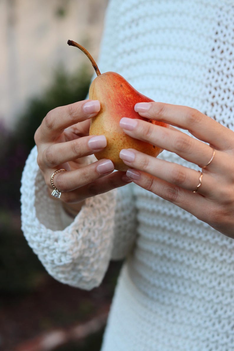May include: A ripe pear with a red and yellow gradient is held by a person. The pear's stem is brown. The person is wearing rings and a white, open-knit sweater. The pear is the focal point of the image.
