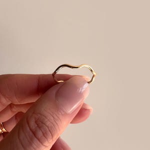 May include: A gold-colored ring with a wavy, organic design. The ring is held up against a neutral background, showcasing its smooth, reflective surface. The ring's unique shape adds a touch of modern elegance to the jewelry.