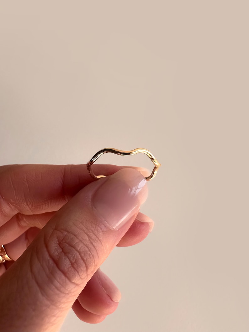 May include: A gold-colored ring with a wavy, organic design. The ring is held up against a neutral background, showcasing its smooth, reflective surface. The ring's unique shape adds a touch of modern elegance to the jewelry.