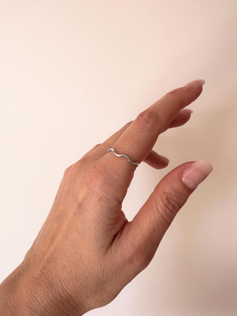 May include: A silver-colored ring with a wavy design is worn on a finger. The ring's design resembles a continuous wave. The background is a plain, light-colored surface.