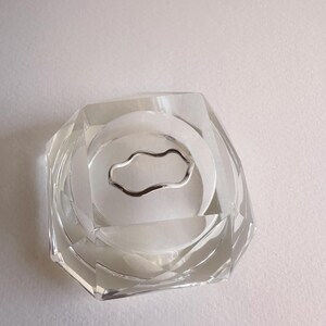 May include: A clear, faceted glass ashtray with a silver-colored, wavy metal ring in the center. The ashtray has a geometric design and is set against a white background. The ashtray is a decorative item.