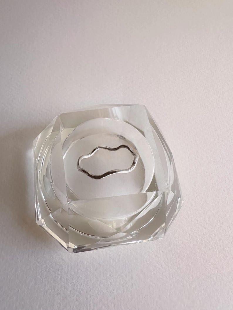 May include: A clear, faceted glass ashtray with a silver-colored, wavy metal ring in the center. The ashtray has a geometric design and is set against a white background. The ashtray is a decorative item.