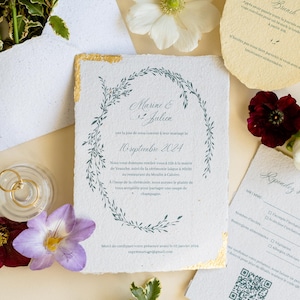 Peut inclure: Faire-part de mariage avec un motif de couronne florale et des détails dorés. L'invitation présente les noms "Marine & Julien" et la date du "10 septembre 2024". Cartes et fleurs supplémentaires.