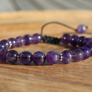 May include: A purple amethyst gemstone bracelet with silver beads and a black cord. The bracelet is adjustable with a sliding knot.