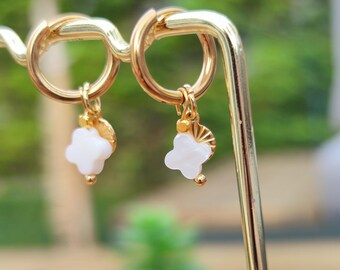 May include: Gold-colored hoop earrings with white clover-shaped charms and small gold beads. The earrings are displayed on a gold stand against a blurred green background, showcasing the jewelry's delicate design.