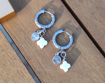 May include: A pair of silver hoop earrings with textured detailing. Each earring features a small, round charm with radial lines and a white clover-shaped charm, creating a delicate and stylish accessory.