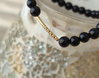 May include: A black beaded bracelet with a gold-colored, geometric-patterned accent. The bracelet is made of glossy, round black beads. The gold accent is a curved bar with a textured surface. The bracelet is displayed on a decorative surface.