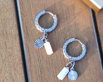 May include: A pair of silver hoop earrings with textured detailing. Each earring features a small white rectangular charm and a silver circular charm with a sunburst design. The earrings are displayed on a wooden surface.