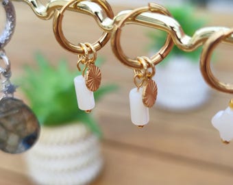 May include: Gold hoop earrings with white rectangular beads and small gold sunburst charms. The earrings are displayed on a gold metal jewelry stand. The background is blurred, with hints of greenery and a wooden surface.