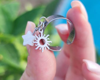 May include: A silver-toned ring with a crescent moon design. Dangling from the moon are a white star and a sun charm. The ring is held up by a person's fingers against a blurred background of green foliage and blue water.