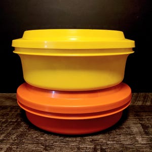 Peut inclure: Deux récipients de conservation des aliments en plastique vintage. Le récipient supérieur est jaune et le récipient inférieur est orange. Les deux récipients ont des couvercles et sont empilés l'un sur l'autre. Les récipients sont posés sur une surface en bois.