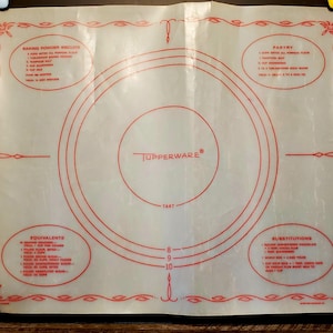 Könnte beinhalten: Eine Tupperware Backmatte mit roten kreisförmigen Führungen und Text. Die Matte enthält Maße für Tortengrößen und Rezepte für Backpulver-Kekse, Gebäck, Äquivalente und Substitutionen. Die Matte hat einen dekorativen roten Rand.