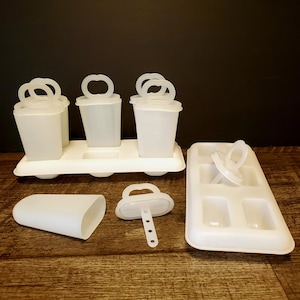 Peut inclure: Ensemble de moules à sucettes glacées en plastique blanc. L'ensemble comprend des moules individuels avec des poignées, un plateau pour les contenir et un moule séparé avec un couvercle. Les articles sont disposés sur une surface en bois.