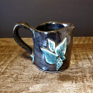May include: A handcrafted ceramic mug with a dark blue glaze and a sculpted leaf design in teal. The mug features a curved handle and a slightly uneven rim, giving it a rustic look. It sits on a wooden surface.