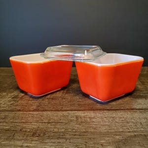 May include: Two square, orange Pyrex dishes with white interiors and clear glass lids. The dishes are sitting on a wooden surface. The lids are clear and square, with a slight lip.