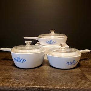 May include: A collection of three white CorningWare casserole dishes with clear glass lids. Each dish has a blue floral pattern. The dishes are displayed on a wooden surface against a dark backdrop. The set includes small, medium, and large dishes.