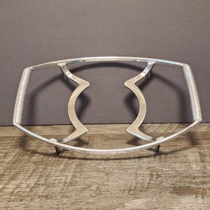 May include: A silver metal hot plate stand with a curved design. The stand has two raised sections in the center and a curved base.