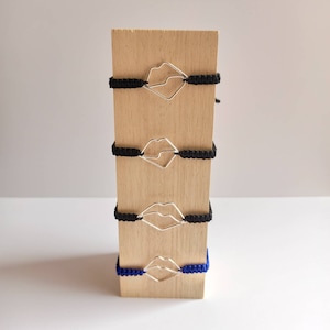 May include: Four friendship bracelets with silver lip charm accents displayed on a wooden stand. Two bracelets are black and two are blue.
