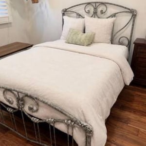 May include: A metal bed frame with a distressed grey finish and ornate scrollwork on the headboard and footboard. The bed is dressed in a solid cream-coloured comforter, two square cream pillows, and a small rectangular light green accent pillow.