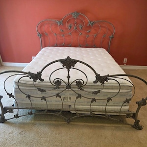 May include: A metal bed frame with a weathered grey finish, featuring an ornate headboard and footboard with intricate scrollwork, floral accents, and curved metal bars. A thick white tufted mattress sits between the head and foot sections.