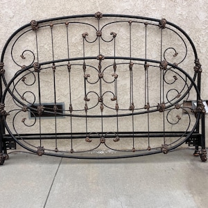 Puede incluir: Un conjunto de cabecero y piecero de cama en metal negro con un diseño arqueado, barras verticales, volutas decorativas y detalles florales en tono bronce en las uniones. La estructura está compuesta por tubos y varillas de metal.