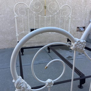 May include: A white metal bed frame featuring a headboard and footboard with intricate scrollwork, floral accents, and a central brass-toned finial. The structure includes a dark brown metal base with adjustable legs for support and stability.