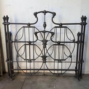 May include: A dark metal bed headboard featuring a symmetrical design with two large circular frames, vertical bars, and decorative finials at the top of the four vertical posts. The structure includes ornate scrollwork and central accents.