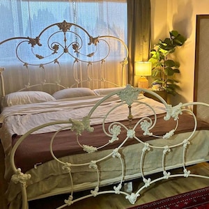 Op de afbeelding: Een wit metalen bedframe met een sierlijk, gebogen hoofd- en voeteneinde met ingewikkeld krulwerk en decoratieve bloemaccenten. Het bed is opgemaakt met lichtgekleurd beddengoed en een donkerbruine sprei als opvallend middelpunt.