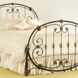 May include: A dark metal bed frame featuring an arched headboard and footboard with intricate scrollwork and gold-toned floral accents. The bed is dressed in a cream-colored comforter with a ruffled bed skirt and several matching pillows.