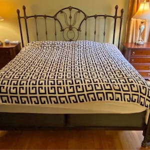 May include: A wrought iron bed frame with a patterned duvet cover. The bed has a decorative headboard and footboard. Two bedside tables with lamps are either side. The room has wooden floors and a window with blinds.