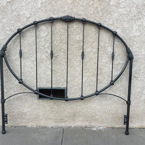 May include: A black metal headboard featuring an oval-shaped frame with vertical spindles. The design includes decorative finials at the connection points and two straight vertical legs with mounting brackets at the base for attachment to a bed frame.