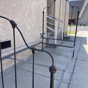 May include: A black metal bed frame featuring a tall headboard and a shorter footboard with arched top rails and decorative finial accents. The frame includes side rails and cross supports, constructed from thin, tubular metal components.