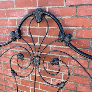 Può includere: Una testiera in metallo nero caratterizzata da una barra superiore curva e complessi motivi a volute. Il design include anelli metallici simmetrici e decorazioni in metallo pressofuso sui giunti, creando una struttura aperta.