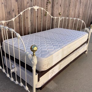Peut inclure: Un lit de repos en métal blanc avec un dossier haut arqué et des rails latéraux ornés de volutes décoratives et de fleurons sphériques en laiton poli. Le lit comprend un matelas blanc matelassé et un cadre de lit gigogne assorti.