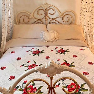 May include: A white bedspread featuring a repeating pattern of embroidered red roses with green leaves, draped over a bed with a cream-coloured metal headboard and footboard. The bed includes two beige pillows and a small heart-shaped decorative pillow.