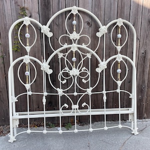 May include: An off-white metal headboard featuring a curved frame with intricate scrollwork and decorative floral accents. The design includes vertical bars with gold-toned metal rods and small, round, patterned ceramic inserts at the center.