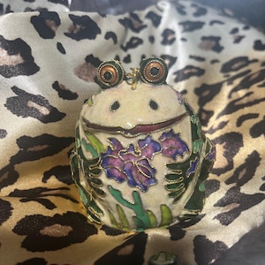 May include: A decorative frog-shaped trinket box. The box is cream-colored with a smiling face, large eyes, and a red mouth. It features purple and blue floral designs and green leaves. The box has gold-colored accents and is set against a leopard print background.