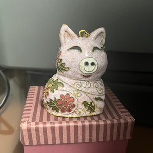 May include: A pink ceramic pig figurine with floral and gold accents. The pig is smiling and has a gold loop for hanging. It sits atop a striped gift box with a matching pink base.