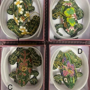 May include: Four decorative frog figurines in individual boxes. Each frog is green with floral and gold accents. The frogs are in various poses and have different floral patterns in white, pink, and red. The boxes are pink with white interiors.