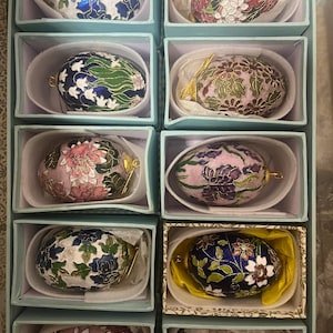 Op de afbeelding: Twaalf decoratieve eieren in individuele dozen. De eieren zijn beschilderd met ingewikkelde bloemmotieven in tinten roze, blauw, groen en wit. De eieren zijn gemaakt met behulp van de cloisonné-techniek, waarbij dunne draden worden gebruikt om compartimenten te creëren die vervolgens worden gevuld met emaille.