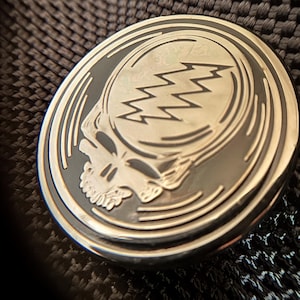 May include: A silver-colored metal pin with a black and white image of a skull with lightning bolts. The skull is surrounded by concentric circles.