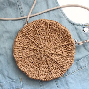 May include: A round straw bag with a light brown woven design and a light gray strap. The bag is on top of a light blue denim fabric.