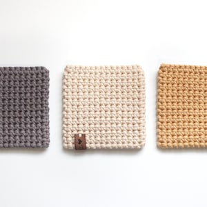 May include: Three crocheted square coasters in gray, white, and beige. Each coaster has a small brown leather tag.