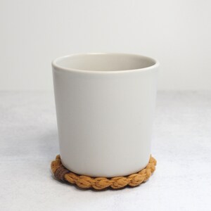 May include: A matte off-white ceramic mug with a smooth, cylindrical body and a wide opening, resting on a circular, braided tan fabric coaster that features a small, dark brown rectangular accent piece on its outer edge.