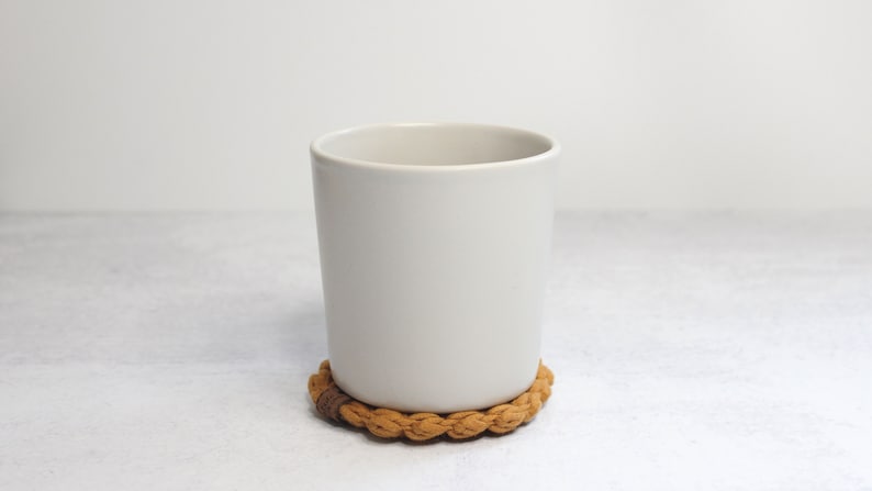 May include: A matte off-white ceramic mug with a smooth, cylindrical body and a wide opening, resting on a circular, braided tan fabric coaster that features a small, dark brown rectangular accent piece on its outer edge.