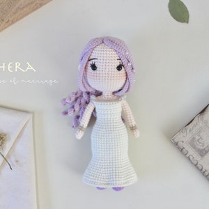 May include: A handmade crochet doll of a woman in a white dress, with lavender hair and accents. The doll has a delicate design, with small details like jewelry and a detailed face. The text "HERA goddess of marriage" is visible.