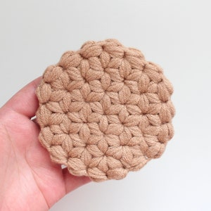 May include: A round, beige crochet coaster featuring a textured, repeating star-like pattern created with thick yarn. The circular item has a scalloped edge and a dense, knotted construction, showcasing a uniform light brown color throughout the piece.