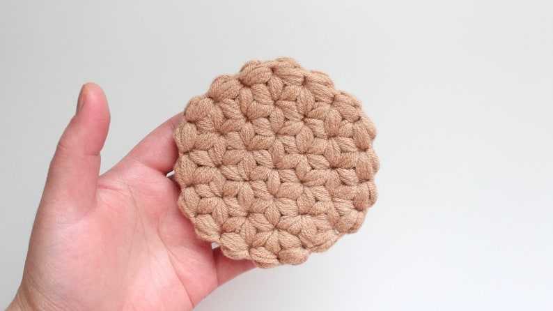 May include: A round, beige crochet coaster featuring a textured, repeating star-like pattern created with thick yarn. The circular item has a scalloped edge and a dense, knotted construction, showcasing a uniform light brown color throughout the piece.