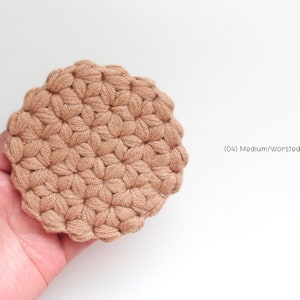 May include: A round, tan-colored crochet coaster featuring a textured puff stitch pattern. The item is labeled with the text (04) Medium/Worsted Cotton Yarn, indicating the material used to create this circular, hand-stitched textile piece.