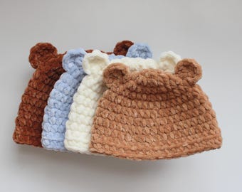 May include: Four crocheted baby hats in shades of brown, light blue, cream, and tan. Each hat has small, rounded bear ears. The hats are made from a soft, textured yarn, perfect for keeping a baby's head warm.